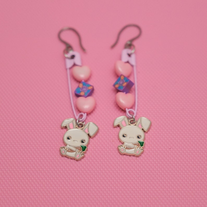 Peut inclure: Une paire de boucles d'oreilles pendantes de couleur rose, violette et blanche. Chaque boucle d'oreille est orn&eacute;e d'un petit charme de lapin blanc, de c&oelig;urs roses et d'un charme carr&eacute; violet.