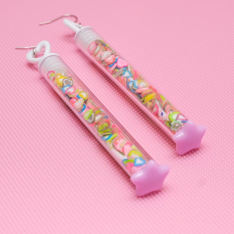 May include: A pair of earrings with clear tubes filled with colorful confetti and a star-shaped charm at the bottom. The earrings are on a pink background.