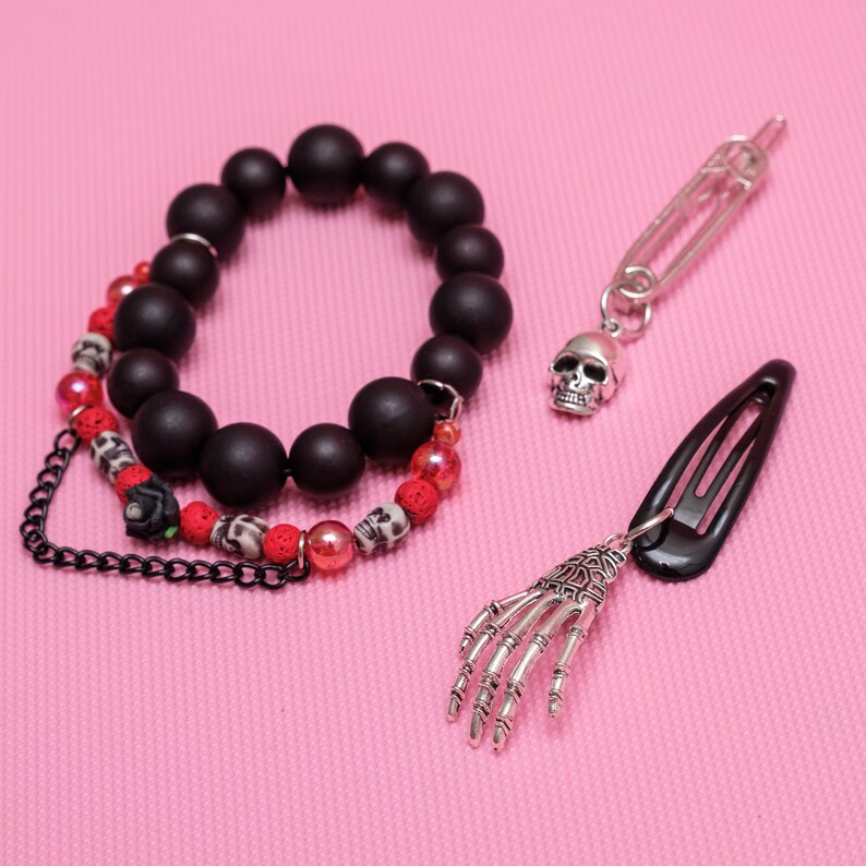 May include: A black beaded bracelet with red and black accents, a silver skull charm on a safety pin, and a black hair clip with a silver skeleton hand charm.