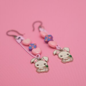 Peut inclure: Une paire de boucles d'oreilles pendantes avec un charme de lapin blanc tenant une carotte verte, des perles de c&oelig;ur roses et des perles &agrave; motifs violets et blancs.