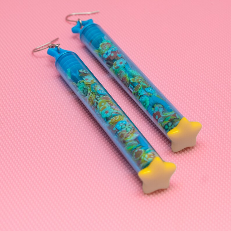 May include: A pair of blue tube earrings with yellow star-shaped tops. The tubes are filled with colorful confetti.