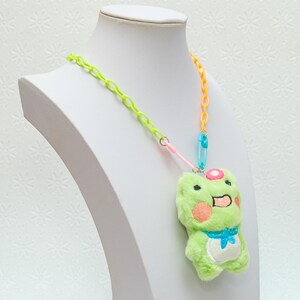 Peut inclure: Un pendentif en forme de grenouille en peluche verte avec une fleur rose sur la t&ecirc;te, suspendu &agrave; un collier en cha&icirc;ne plastique multicolore. Le collier est compos&eacute; de maillons en plastique vert lime, orange et bleu.