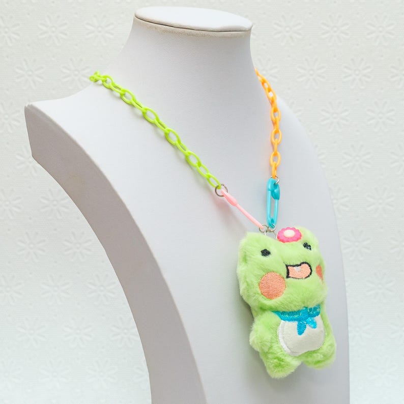Peut inclure: Un pendentif en forme de grenouille en peluche verte avec une fleur rose sur la t&ecirc;te, suspendu &agrave; un collier en cha&icirc;ne plastique multicolore. Le collier est compos&eacute; de maillons en plastique vert lime, orange et bleu.