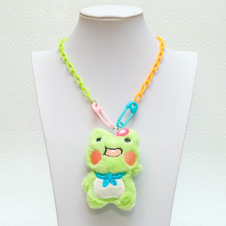 May include: A green frog plush toy with a pink flower on its head is attached to a multi-coloured plastic chain necklace with safety pins. The necklace is yellow, green, and blue.