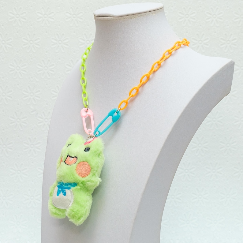 Peut inclure: Un collier en cha&icirc;ne de plastique multicolore avec un pendentif en forme de grenouille en peluche verte. La cha&icirc;ne est compos&eacute;e de maillons en plastique vert lime, orange et bleu. Le collier est s&eacute;curis&eacute; par deux &eacute;pingles de s&ucirc;ret&eacute; roses.
