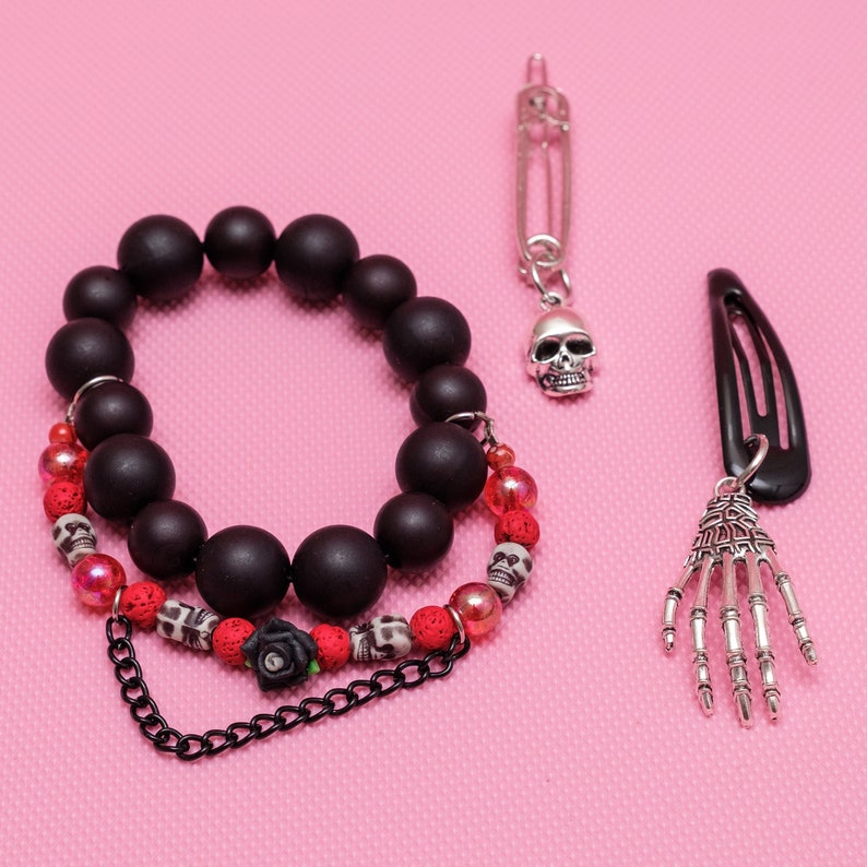 May include: A black beaded bracelet with red and black skull beads, a black hair clip with a silver skeleton hand charm, and a silver safety pin with a silver skull charm.