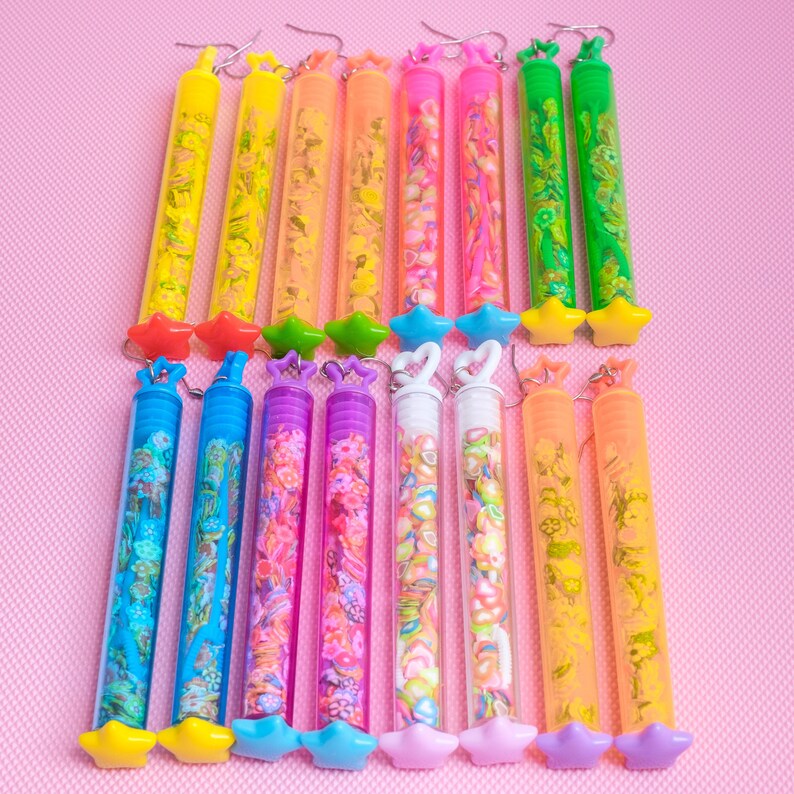 May include: A set of 12 pairs of colorful tube earrings. Each tube is filled with colorful confetti and has a star-shaped charm at the bottom. The earrings are arranged in two rows on a pink background.