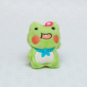 Peut inclure: Un jouet en peluche de grenouille verte avec une fleur rose sur la t&ecirc;te. Il a un n&oelig;ud papillon bleu et un ventre blanc.
