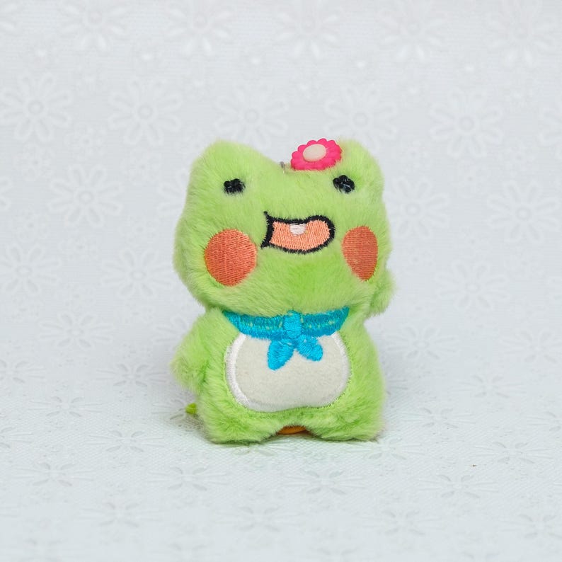 Peut inclure: Un jouet en peluche de grenouille verte avec une fleur rose sur la t&ecirc;te. Il a un n&oelig;ud papillon bleu et un ventre blanc.