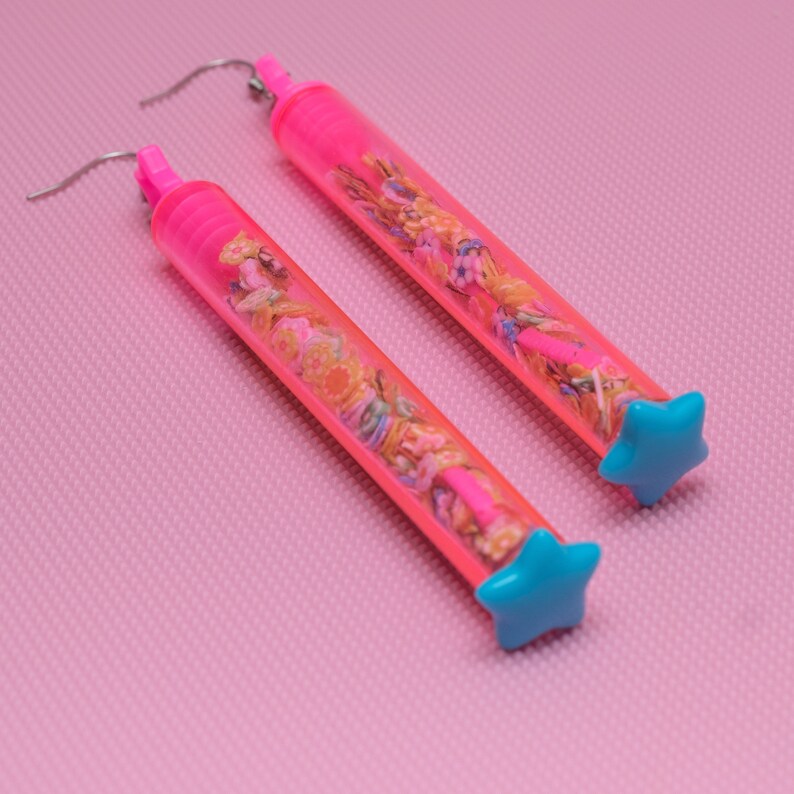 May include: A pair of pink earrings with a clear tube filled with colorful confetti and a blue star charm at the end.