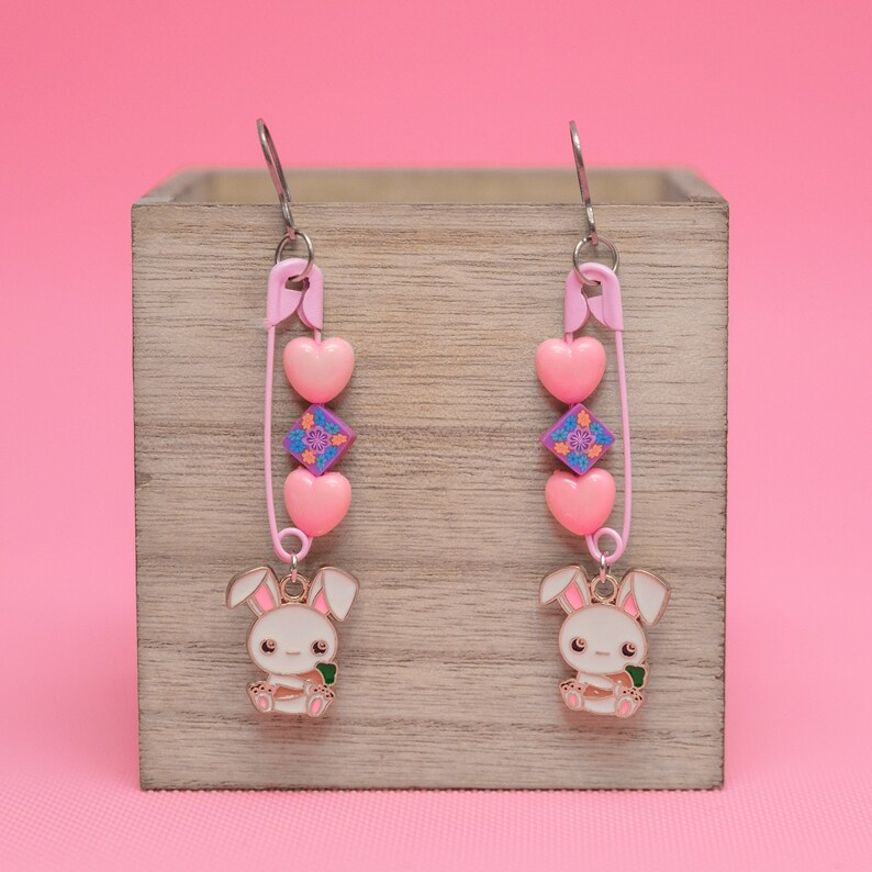 Peut inclure: Une paire de boucles d'oreilles pendantes roses et blanches avec un charme de broche de s&ucirc;ret&eacute;, des c&oelig;urs roses, un charme floral violet et rose et un charme de lapin blanc tenant une carotte.