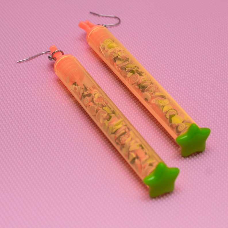 May include: A pair of orange tube earrings with green star-shaped tops. The tubes are filled with colorful confetti.