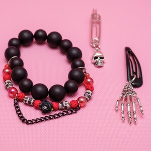 May include: A black beaded bracelet with red and white beads, a black chain, and a black rose charm. The bracelet is paired with a silver skull charm on a safety pin and a silver skeleton hand charm on a black hair clip.