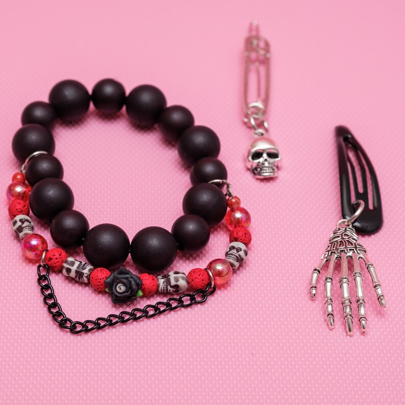 May include: A black beaded bracelet with red and white beads, a black chain, and a black rose charm. The bracelet is paired with a silver skull charm on a safety pin and a silver skeleton hand charm on a black hair clip.