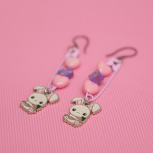 Peut inclure: Une paire de boucles d'oreilles pendantes avec des perles roses et violettes, des c&oelig;urs blancs et un charme de lapin. Les boucles d'oreilles sont sur un fond rose.