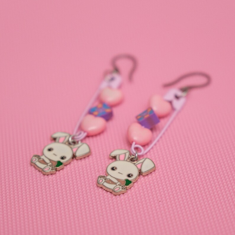 Peut inclure: Une paire de boucles d'oreilles pendantes avec des perles roses et violettes, des c&oelig;urs blancs et un charme de lapin. Les boucles d'oreilles sont sur un fond rose.