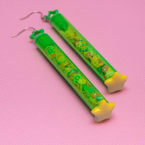May include: A pair of green tube earrings with yellow star charms. The tubes are filled with small green and yellow flowers.