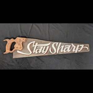 以下が含まれることがあります： 木製のハンドルと金属製の刃を持つヴィンテージの手のこぎり。刃には「Stay Sharp」という文字が白く塗られています。