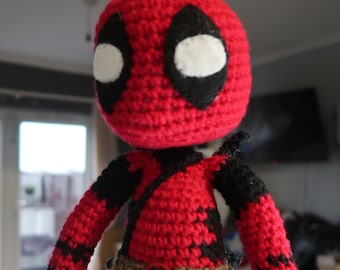 Puede incluir: Un personaje de Deadpool de crochet rojo y negro con un cinturón marrón. El personaje tiene un ojo blanco con un contorno negro.