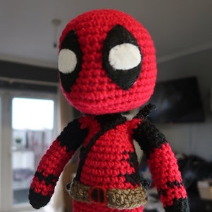 Op de afbeelding: Een rood en zwart gebreid Deadpool-personage met een bruine riem. Het personage heeft een wit oog met een zwarte omtrek.