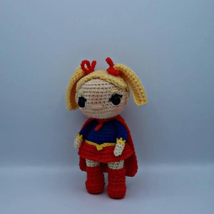 Op de afbeelding: Een gehaakte pop met blond haar, rode strikjes en een rood en blauw superheldenpak.
