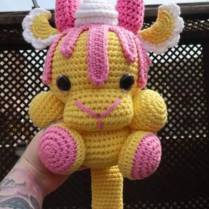 Op de afbeelding: Een gehaakt knuffeldier met een geel lichaam, roze hoorns en een wit, pluche hoofd. Het dier heeft roze voeten en een lange roze staart.