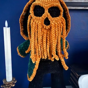 Peut inclure: Une poupée en crochet peluche avec une cape à capuche vert foncé et un visage texturé jaune moutarde doté de longs tentacules pendants. La figurine présente de grandes orbites vides et une petite bouche, faite de fil doux et duveteux.