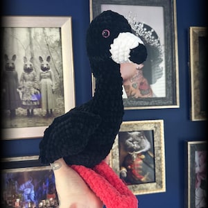 May include: A handmade black plush goose with a white beak and red feet. The plush toy has pink eyes and is made of soft, textured yarn. The goose is a whimsical, decorative item.