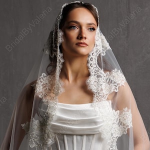 May include: A white wedding dress with a corset bodice and a flowing skirt. The dress is paired with a sheer veil trimmed with delicate lace. The image is set against a neutral gray backdrop.