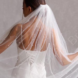 May include: A white bridal veil adorned with small, scattered pearls. The sheer fabric falls over a white wedding dress, creating a soft, elegant appearance. The veil is held out, displaying its delicate design and texture.