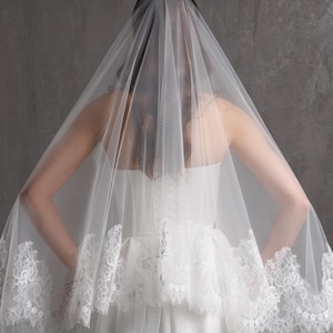 Puede incluir: Un velo de novia blanco con delicados bordes de encaje. El velo est&aacute; drapeado sobre un vestido de novia blanco, creando un aspecto suave y rom&aacute;ntico. Los detalles de encaje a&ntilde;aden un toque de elegancia al dise&ntilde;o.