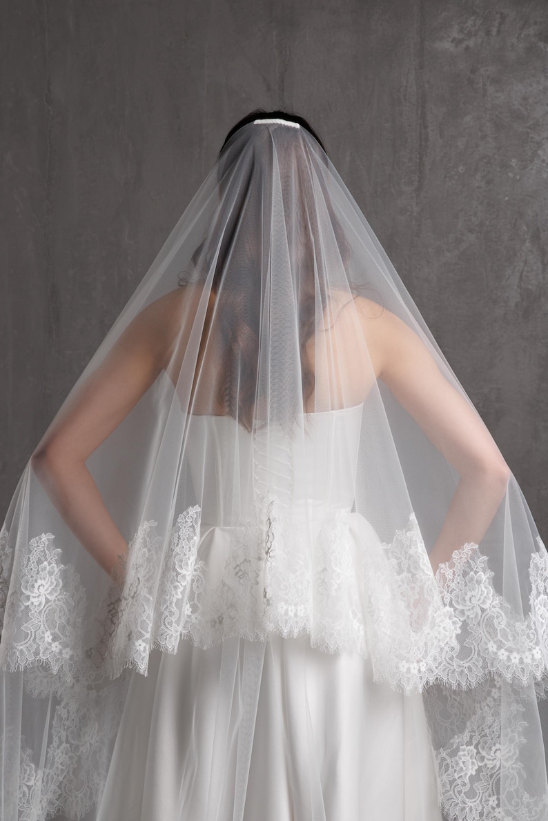 Puede incluir: Un velo de novia blanco con delicados bordes de encaje. El velo est&aacute; drapeado sobre un vestido de novia blanco, creando un aspecto suave y rom&aacute;ntico. Los detalles de encaje a&ntilde;aden un toque de elegancia al dise&ntilde;o.