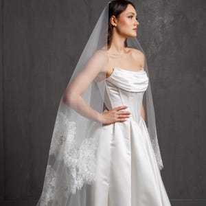 Puede incluir: Vestido de novia de sat&eacute;n blanco con corpi&ntilde;o sin tirantes y falda amplia. Un velo transparente con ribete de encaje cae por la espalda. La novia lleva un anillo.