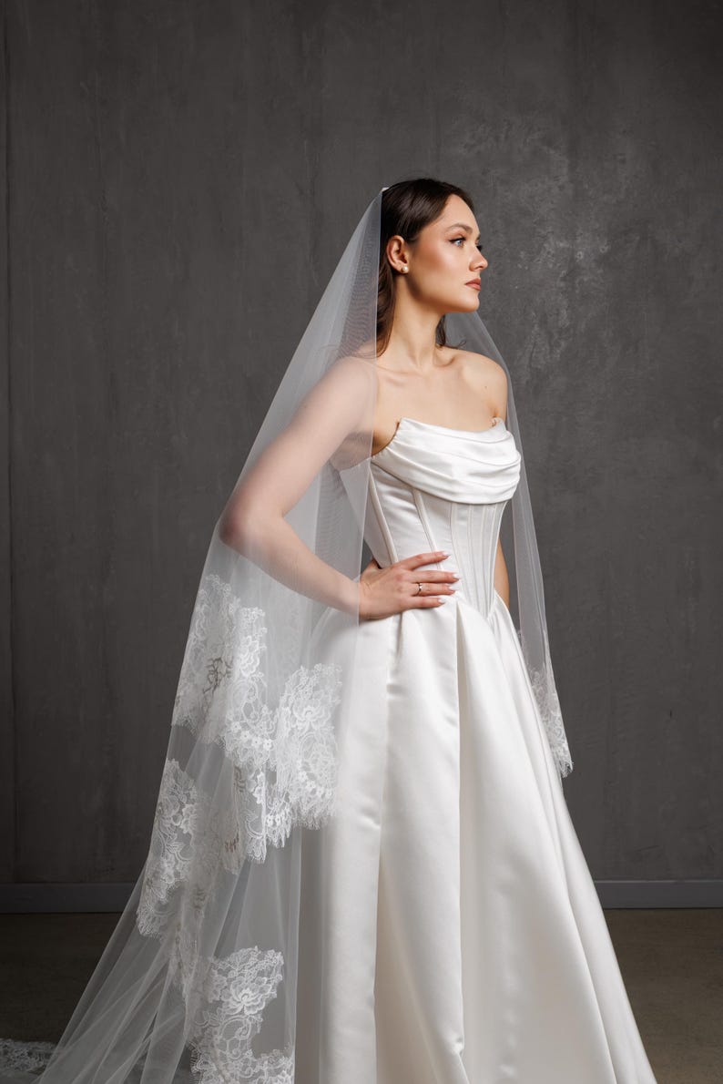 Puede incluir: Vestido de novia de sat&eacute;n blanco con corpi&ntilde;o sin tirantes y falda amplia. Un velo transparente con ribete de encaje cae por la espalda. La novia lleva un anillo.