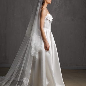 Puede incluir: Un vestido de novia blanco sin tirantes con una falda amplia y un velo largo y transparente. El velo presenta delicados detalles de encaje en los bordes. El vestido tiene un corpi&ntilde;o ajustado y una falda fluida.