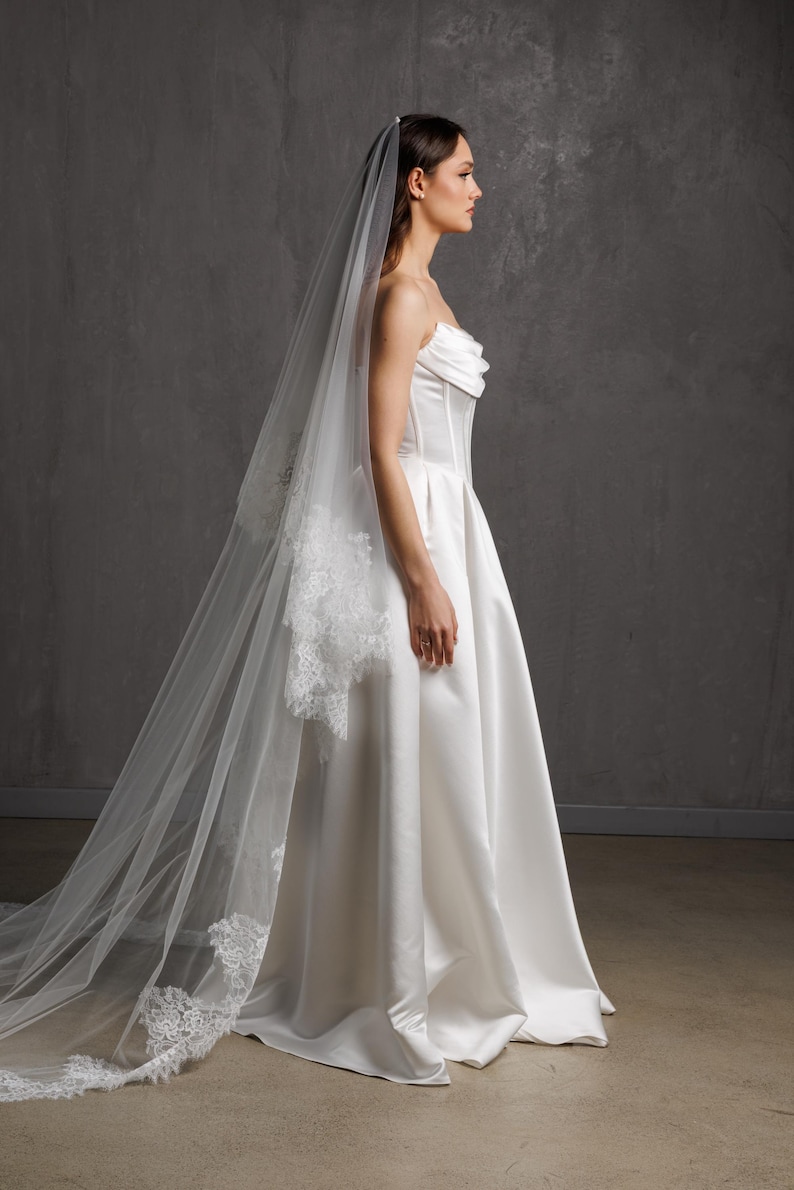 Puede incluir: Un vestido de novia blanco sin tirantes con una falda amplia y un velo largo y transparente. El velo presenta delicados detalles de encaje en los bordes. El vestido tiene un corpi&ntilde;o ajustado y una falda fluida.
