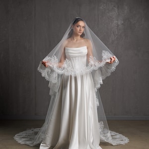Puede incluir: Un vestido de novia de sat&eacute;n blanco con un corpi&ntilde;o sin tirantes y una falda fluida. El vestido se combina con un velo transparente adornado con delicado encaje. El velo cae por la espalda, creando un look nupcial cl&aacute;sico.
