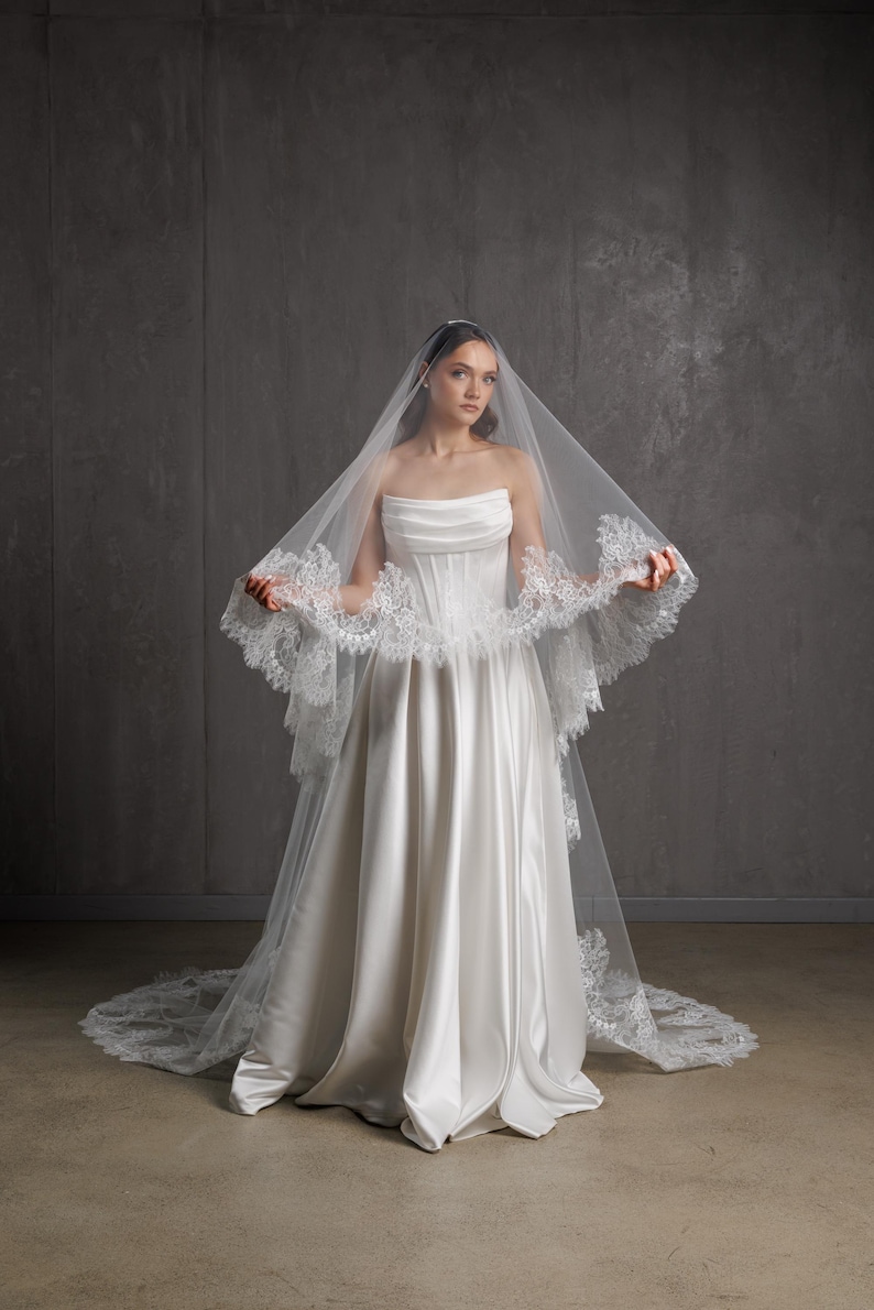 Puede incluir: Un vestido de novia de sat&eacute;n blanco con un corpi&ntilde;o sin tirantes y una falda fluida. El vestido se combina con un velo transparente adornado con delicado encaje. El velo cae por la espalda, creando un look nupcial cl&aacute;sico.