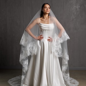 Puede incluir: Vestido de novia de sat&eacute;n blanco con corpi&ntilde;o sin tirantes y falda amplia. Un velo transparente con ribete de encaje cubre los hombros. La novia posa con las manos en las caderas frente a un fondo gris.