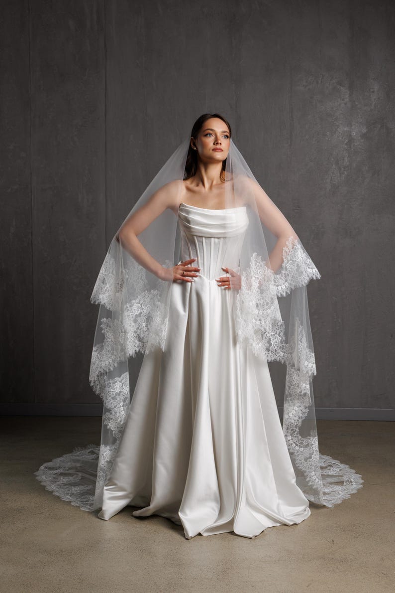 Puede incluir: Vestido de novia de sat&eacute;n blanco con corpi&ntilde;o sin tirantes y falda amplia. Un velo transparente con ribete de encaje cubre los hombros. La novia posa con las manos en las caderas frente a un fondo gris.