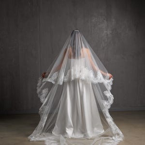 Puede incluir: Un vestido de novia blanco con un velo largo y fluido. El velo est&aacute; adornado con encaje delicado y cae sobre los hombros y la espalda del vestido. El vestido tiene un dise&ntilde;o simple y elegante.