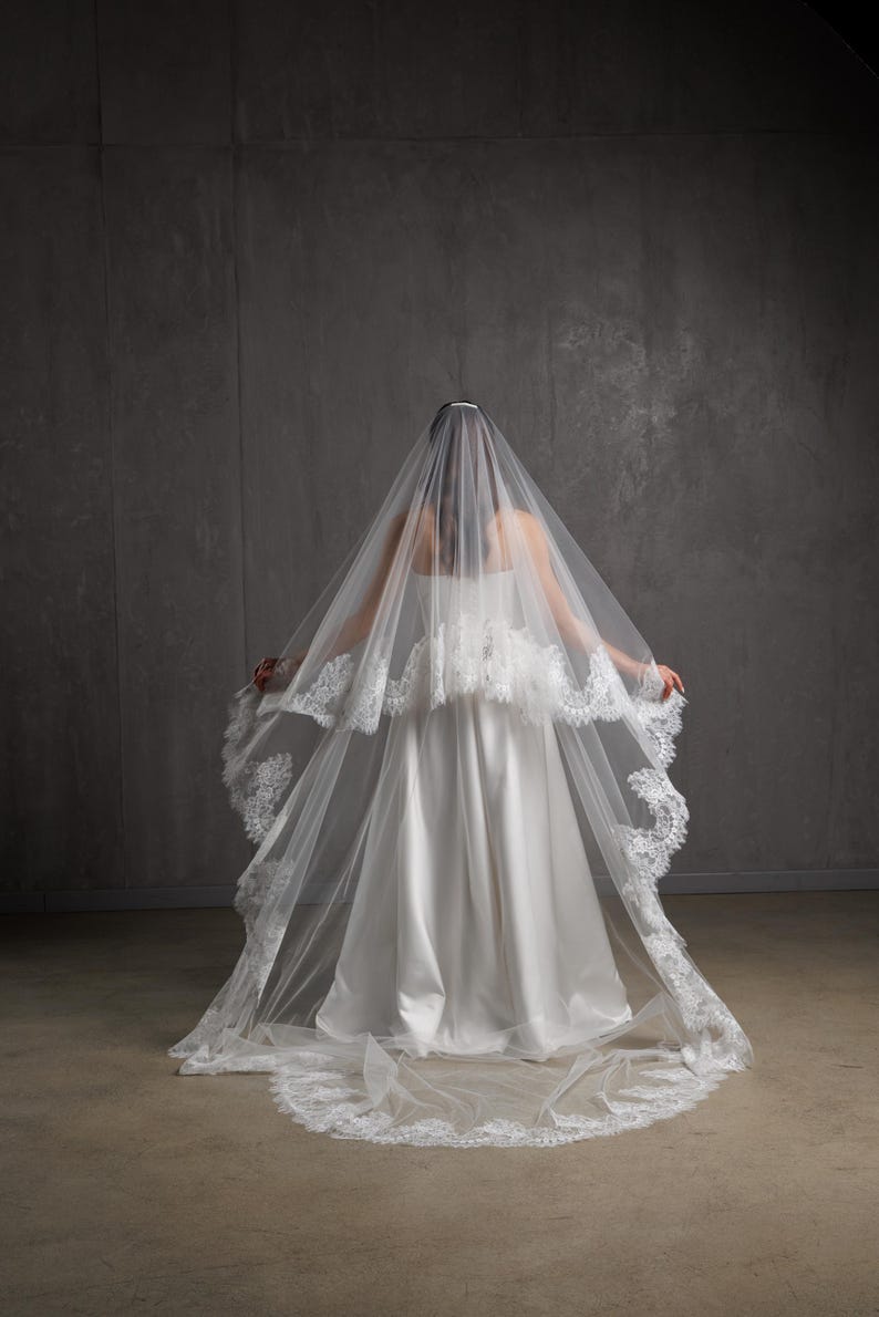 Puede incluir: Un vestido de novia blanco con un velo largo y fluido. El velo est&aacute; adornado con encaje delicado y cae sobre los hombros y la espalda del vestido. El vestido tiene un dise&ntilde;o simple y elegante.