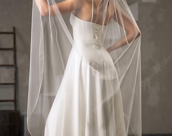 May include: A bride in a white wedding dress with a spaghetti strap bodice and a flowing skirt. She is holding a sheer veil that cascades down her back. The dress has a corset back. The background is a neutral gray.