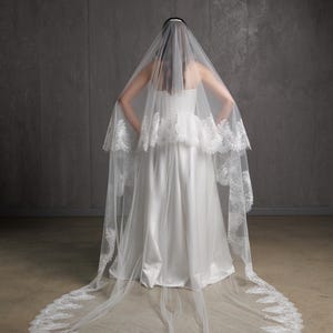 Puede incluir: Un velo de novia blanco con un delicado ribete de encaje. El velo es largo y fluido, con un borde redondeado. La novia lleva un vestido de novia blanco sin tirantes. El fondo es gris neutro.