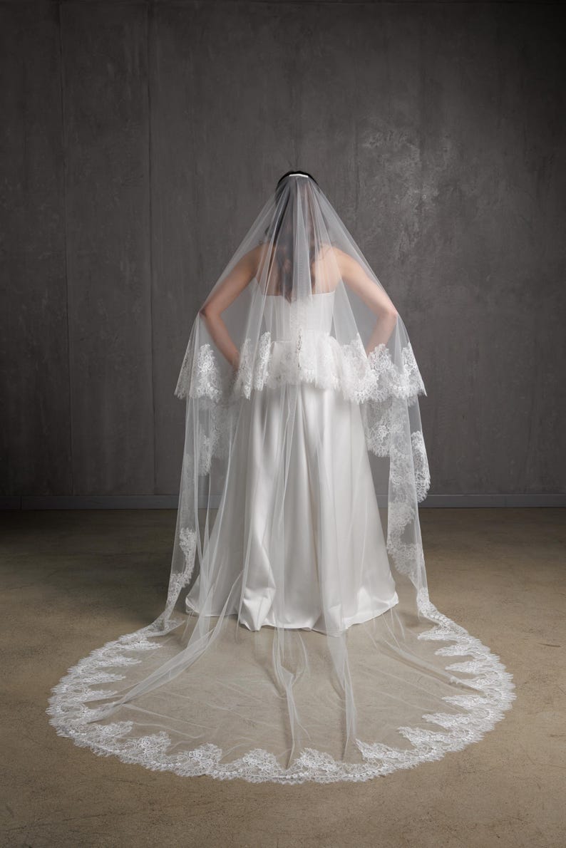 Puede incluir: Un velo de novia blanco con un delicado ribete de encaje. El velo es largo y fluido, con un borde redondeado. La novia lleva un vestido de novia blanco sin tirantes. El fondo es gris neutro.