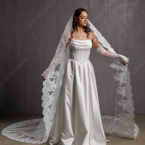 May include: A white strapless wedding gown with a corset bodice and a flowing skirt. The bride is wearing a long, lace-trimmed veil and matching lace gloves. The dress is floor-length and made of a satin-like material.
