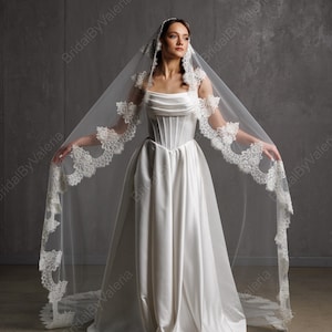 May include: A white satin wedding dress with a corset bodice and a flowing skirt. The bride is wearing a long, sheer veil with delicate lace trim. The veil is spread out, showcasing the intricate details and design.