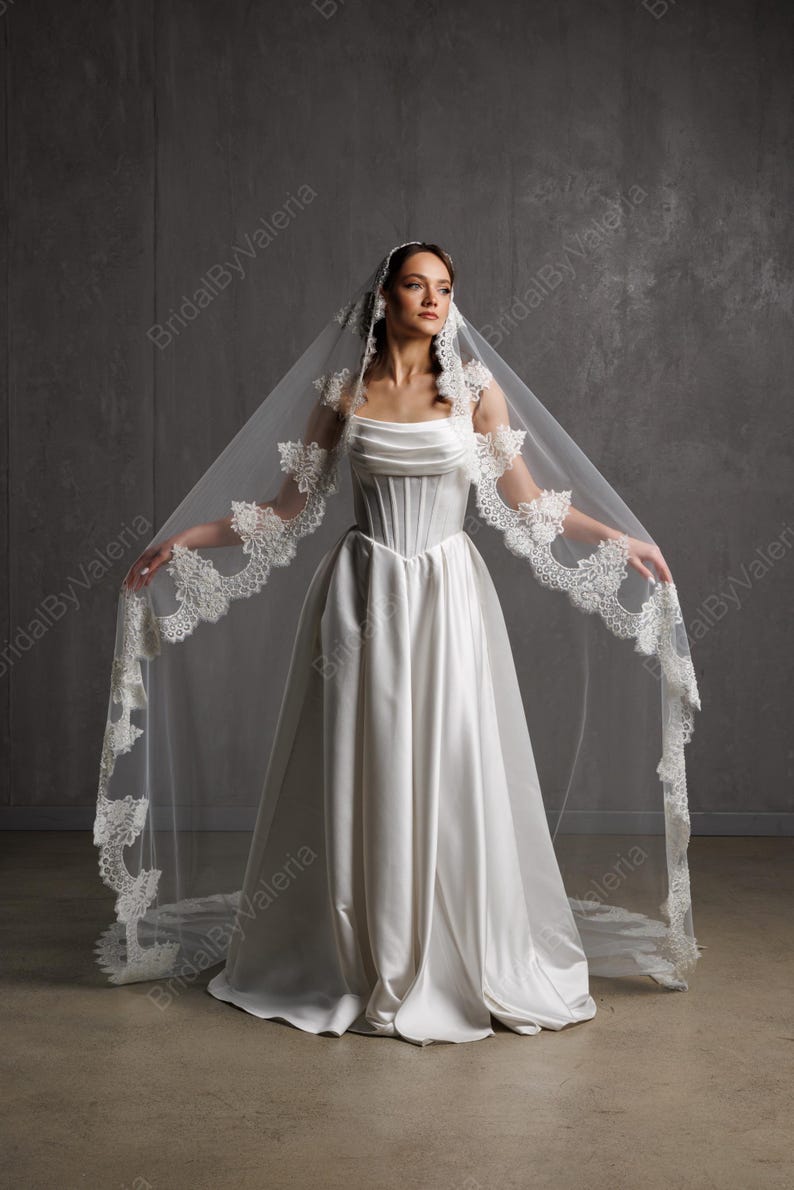 May include: A white satin wedding dress with a corset bodice and a flowing skirt. The bride is wearing a long, sheer veil with delicate lace trim. The veil is spread out, showcasing the intricate details and design.