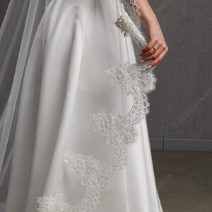 May include: A white satin wedding dress with a flowing skirt and a delicate lace trim. The dress features a long, sheer veil and intricate lace detailing on the sleeve. The overall design is elegant and classic.