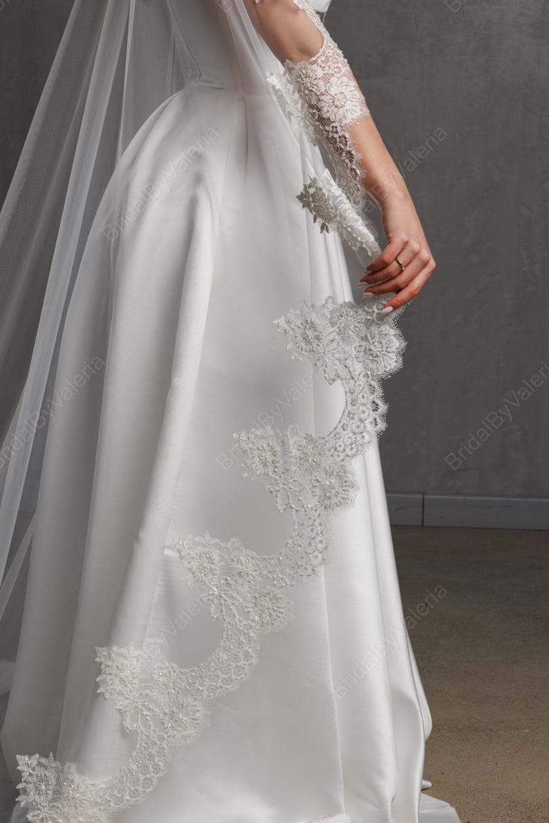 May include: A white satin wedding dress with a flowing skirt and a delicate lace trim. The dress features a long, sheer veil and intricate lace detailing on the sleeve. The overall design is elegant and classic.